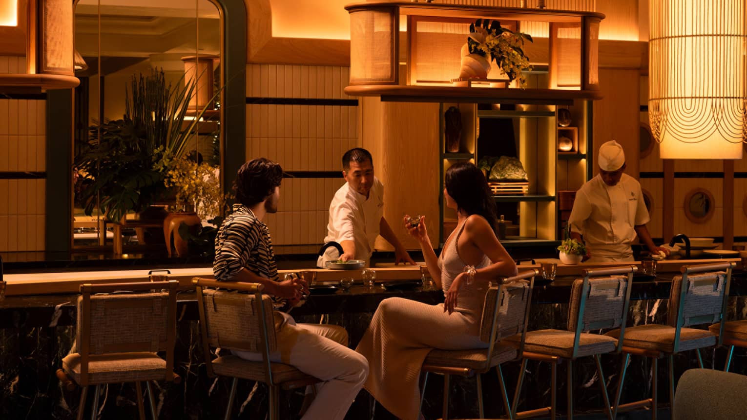 Two people sit at glamorous sushi counter at luxury hotel, dimly lit
