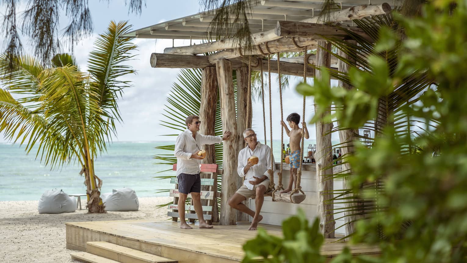 Two men smile at a boy standing on a beach swing at Ile Aux Cerfs