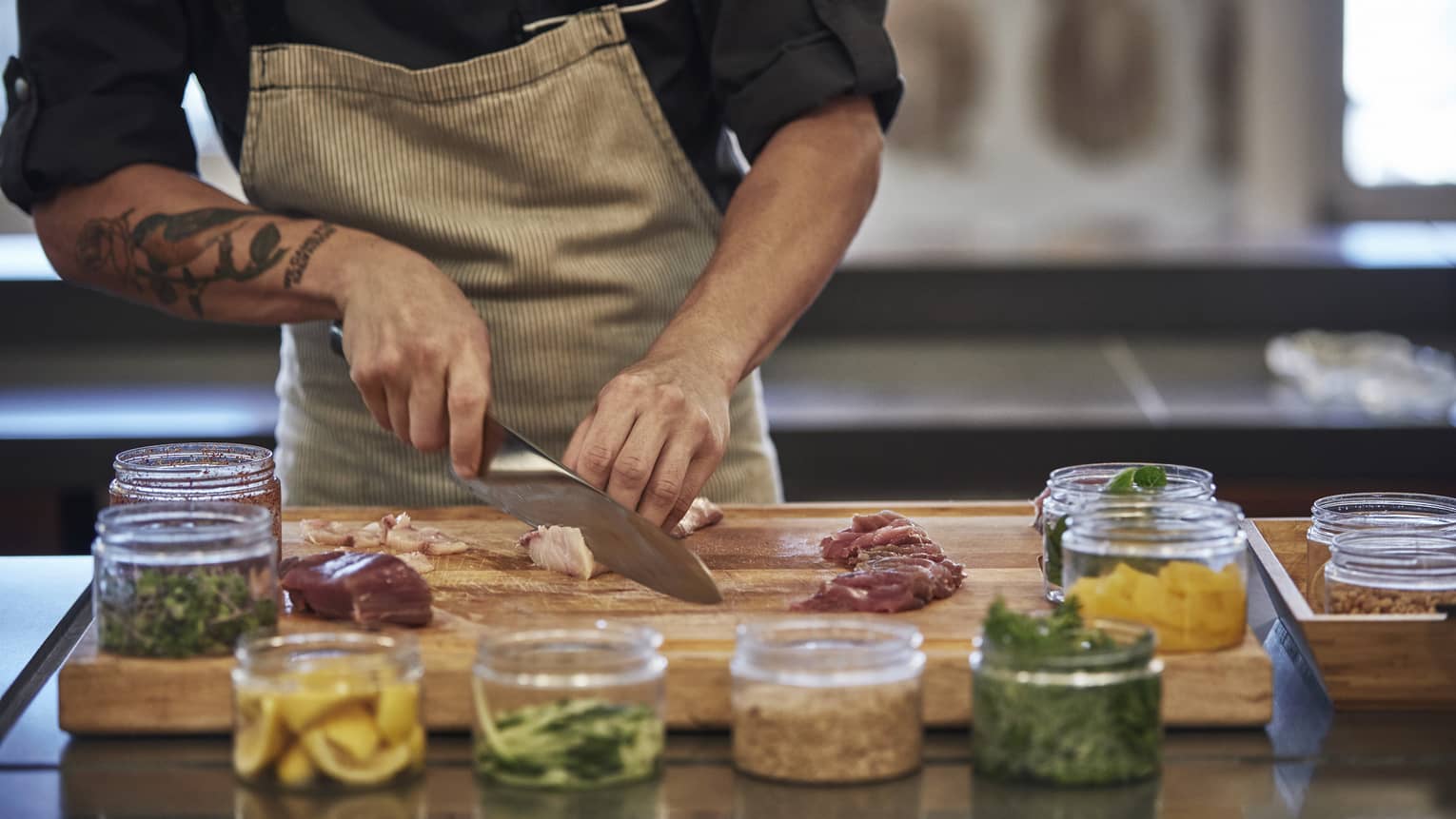 Close-up of chef slicing raw fish on wood butcher's block by glass jars with garnishes 