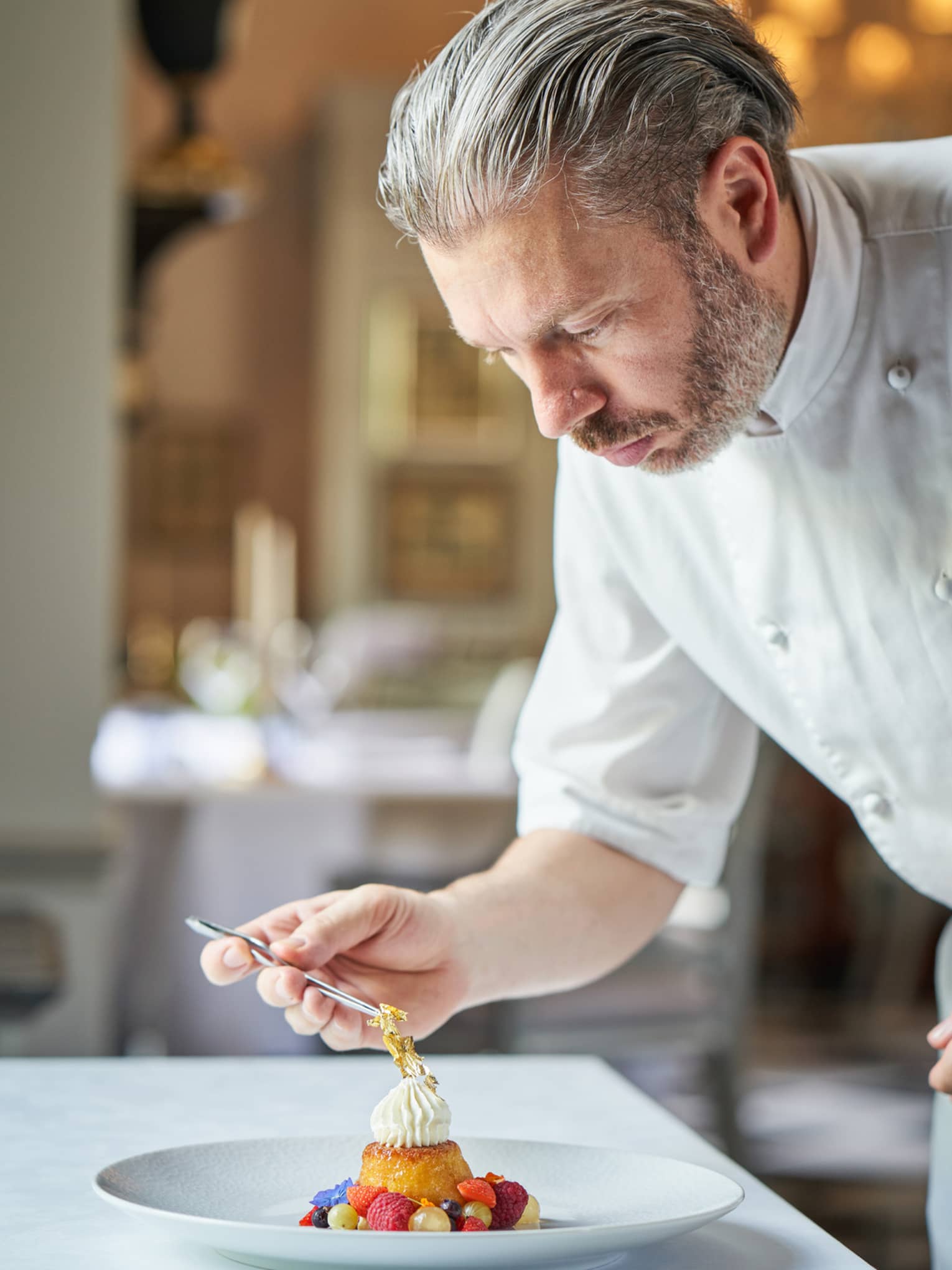 Young grey-haired chef in white chef’s coat delicately placing a garnish atop a dessert on white plate