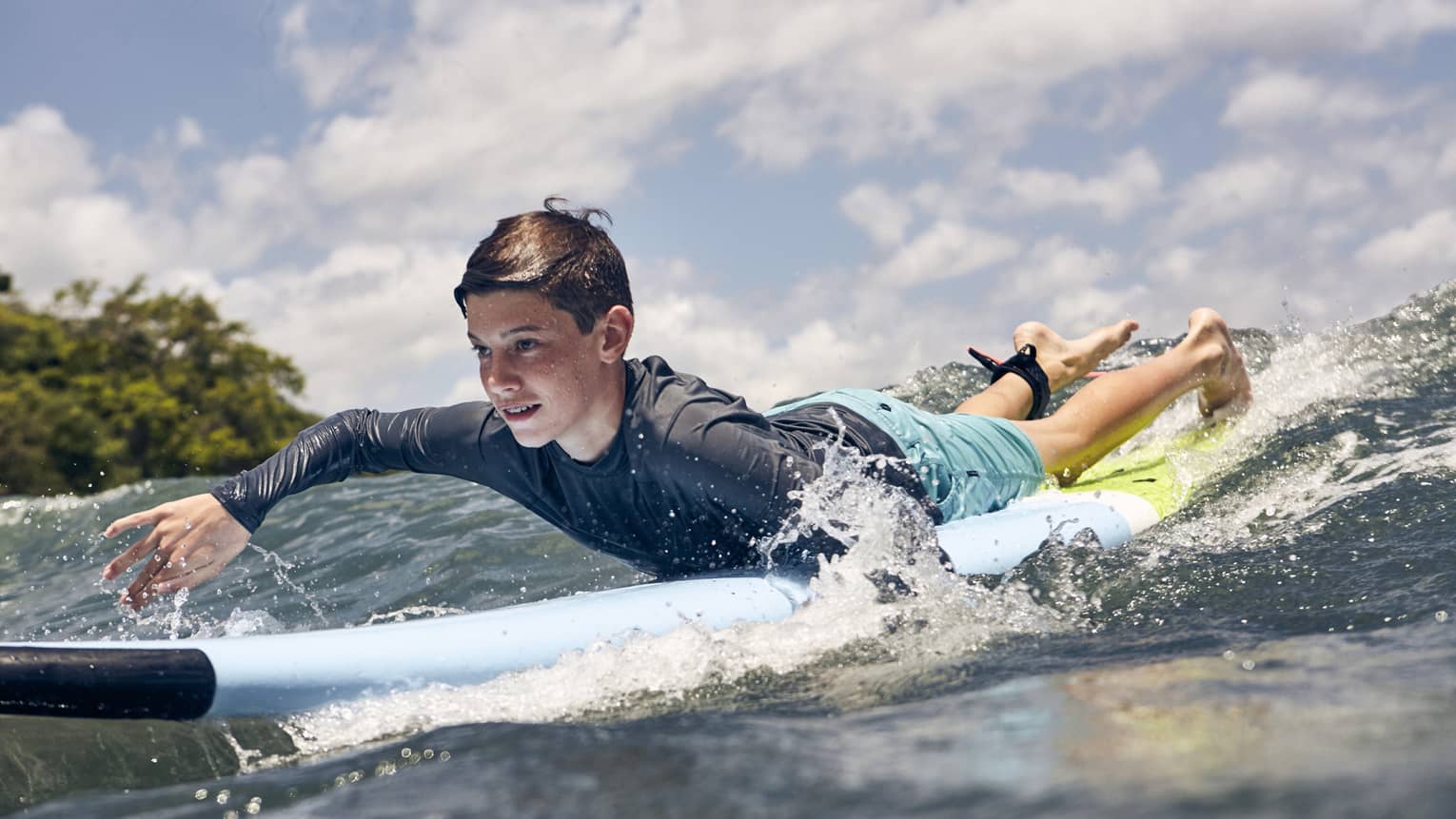 Close-up of a child paddling a surfboard over a wave in the glistening sea, water splashing around the sides of the board.