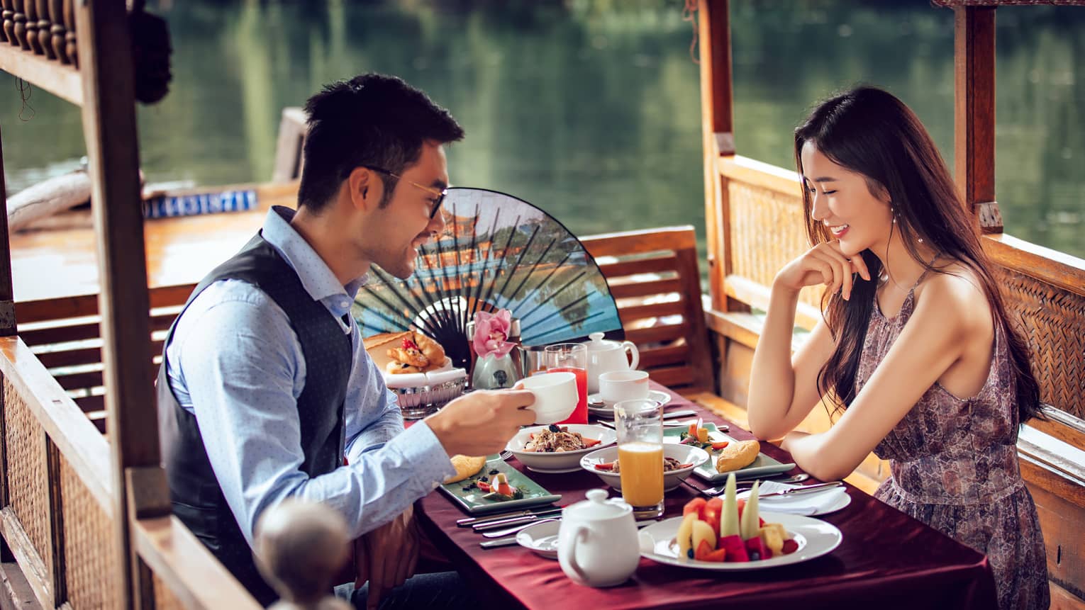 Smiling couple eats brunch at small wood covered table by water, colourful paper fan