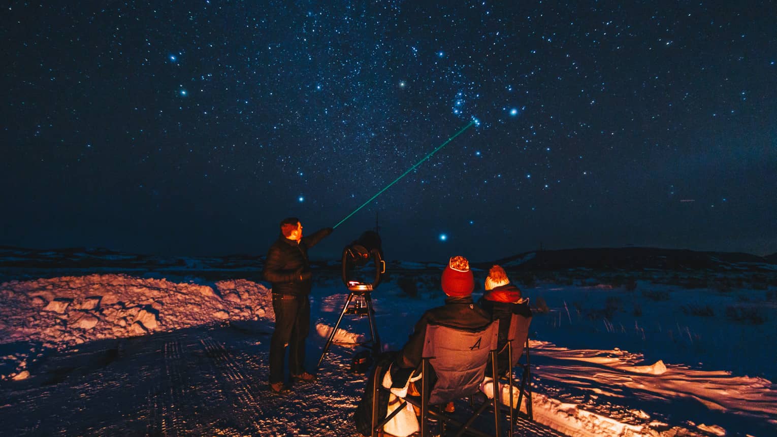 Group of people stargazing on a snowy night, with one person pointing a green laser at the sky, surrounded by chairs and a telescope 