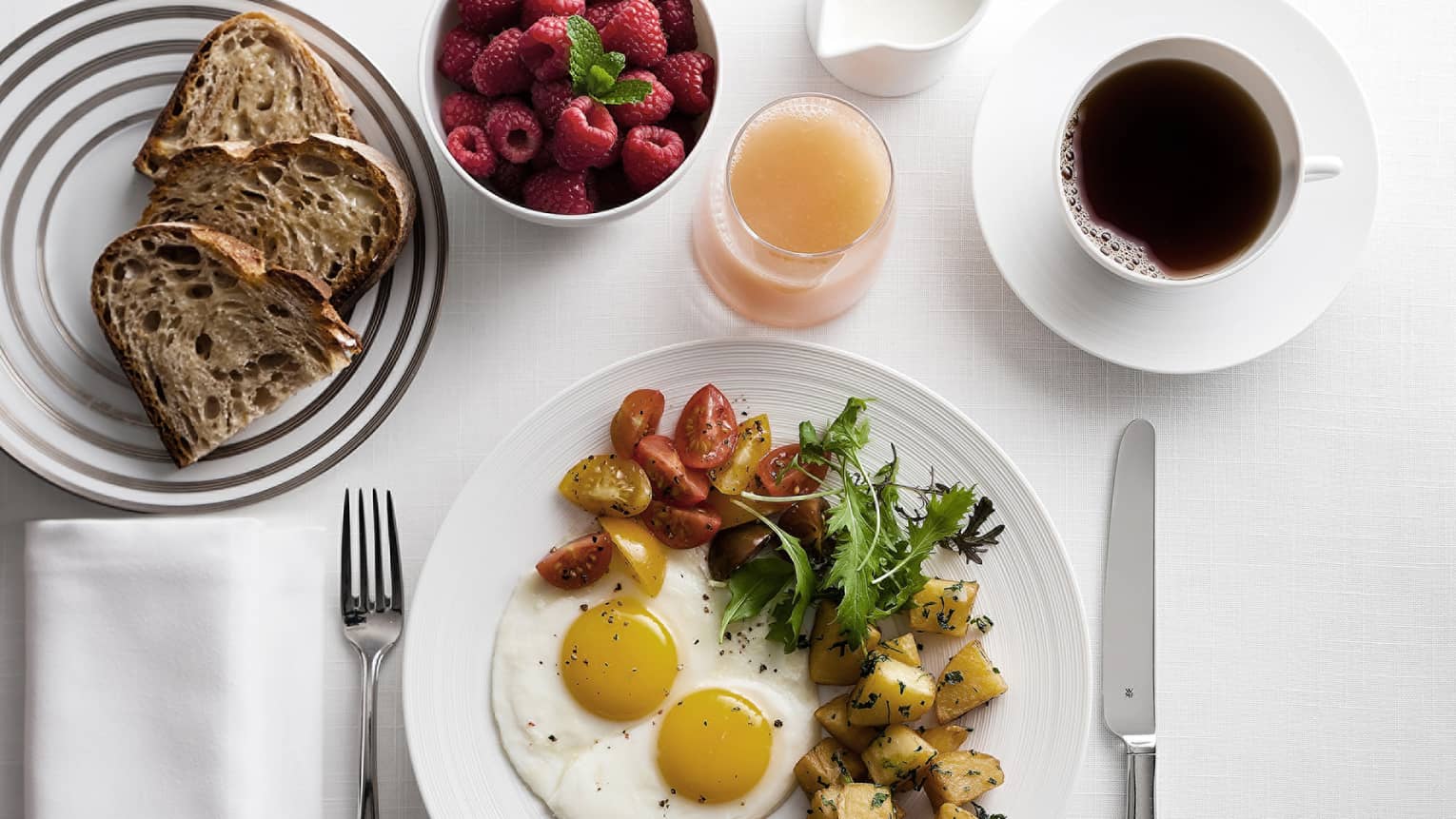 Breakfast plate with sunny-side-up eggs, cherry tomatoes, greens, and roasted potatoes, alongside toast, raspberries, grapefruit juice and coffee.
