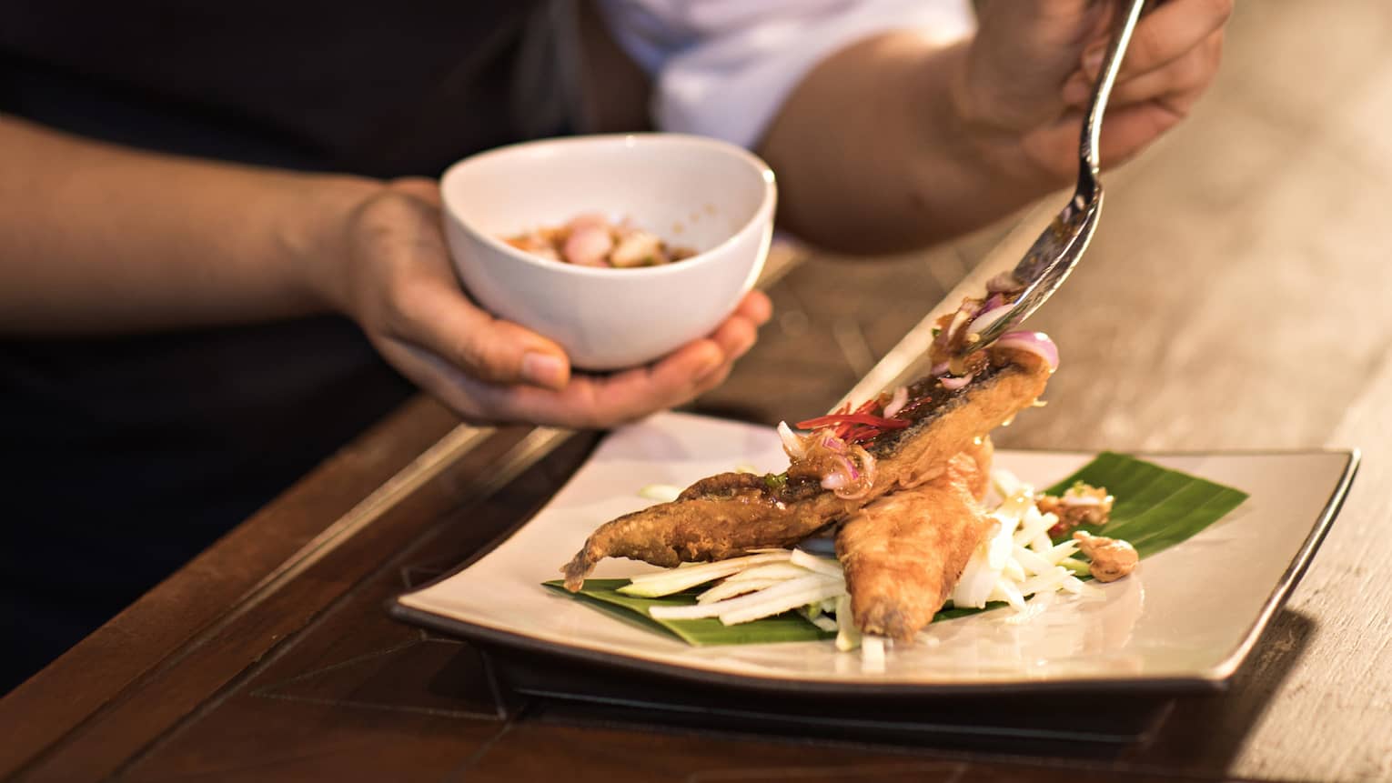 Woman spoons sauce onto Pra Ram Long Song fried fish on plate