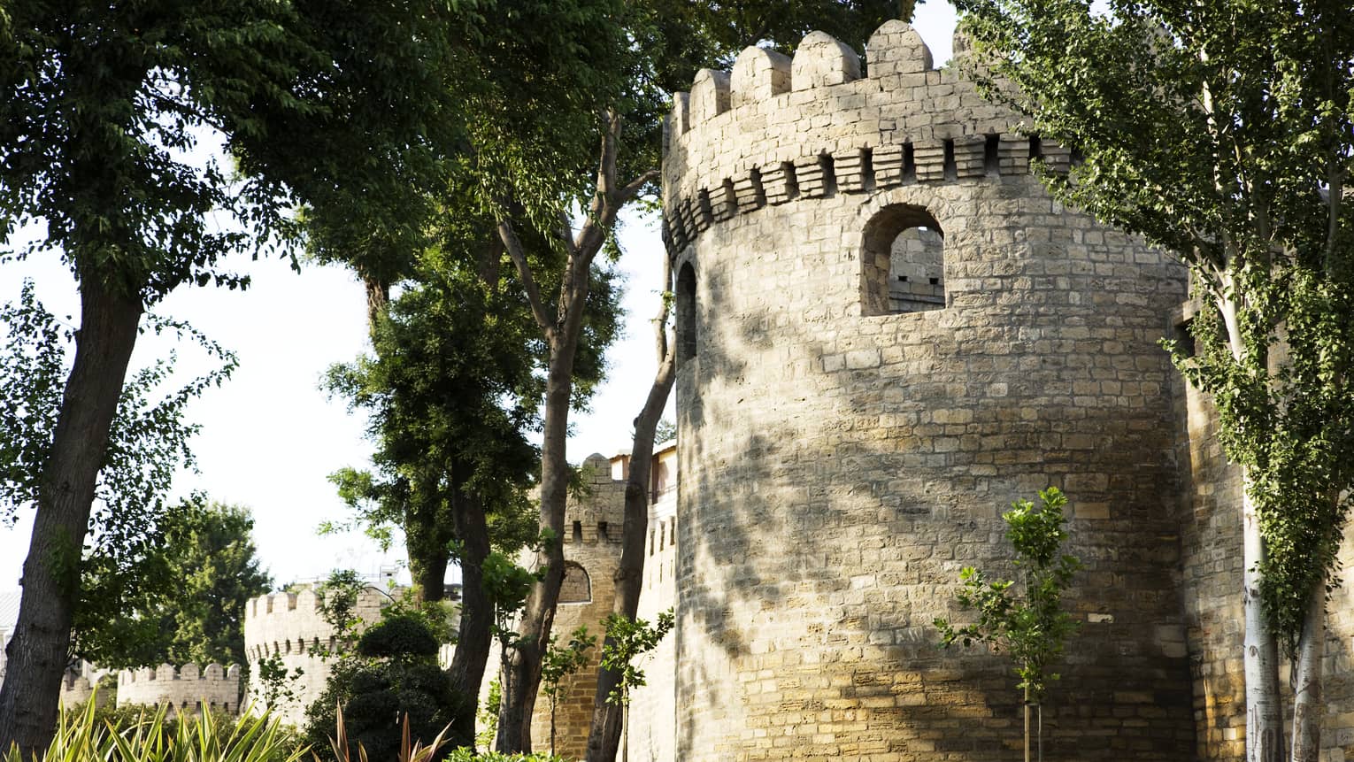A cylindrical structure marked by loophole openings and grey stone exterior: part of a crenelated tower surrounded by trees. 