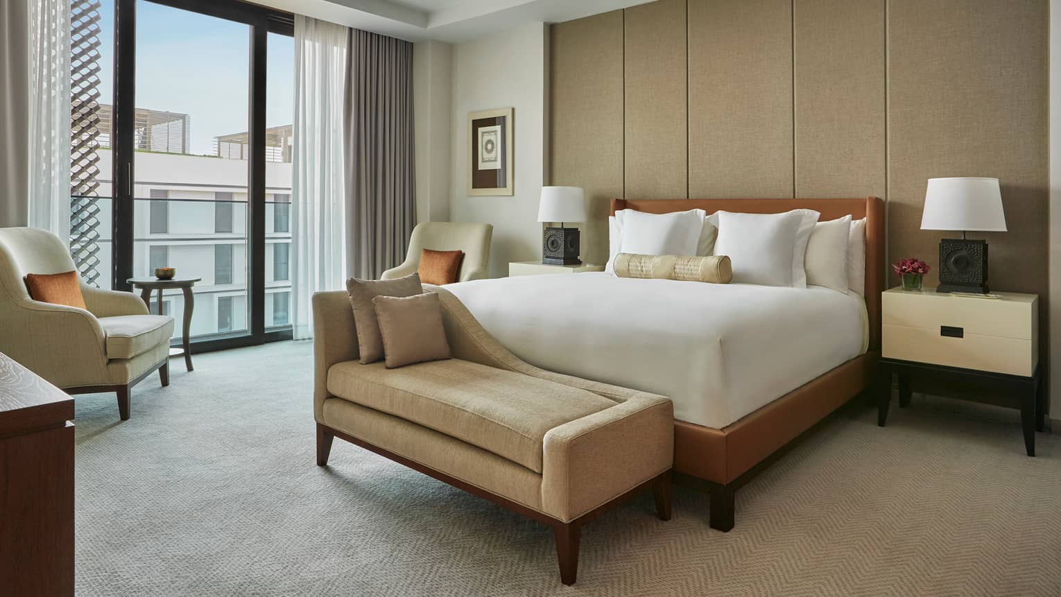 Premier Suite bed with small beige chaise at foot, modern armchairs by large window