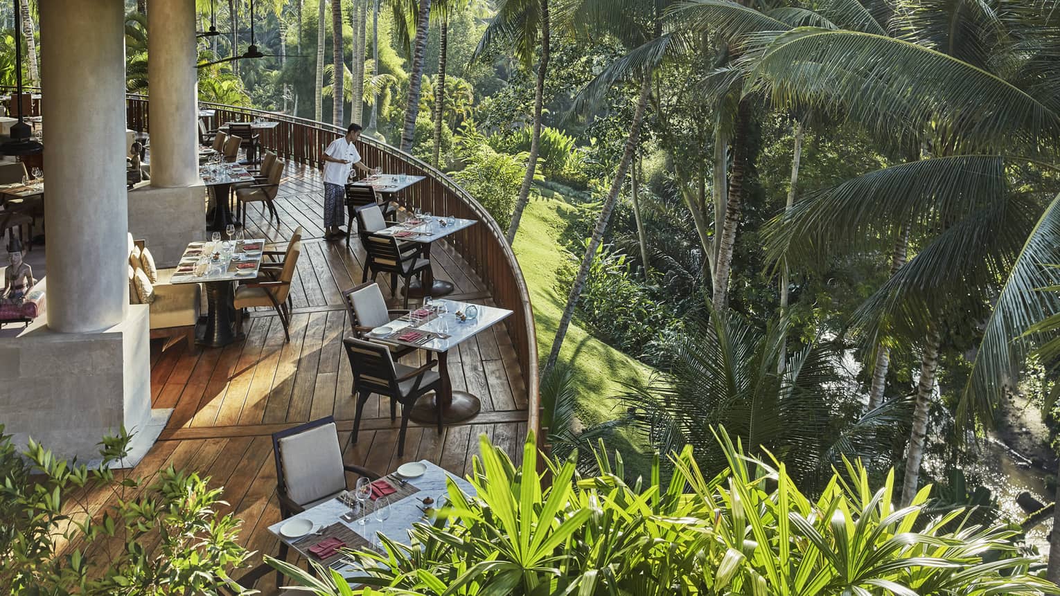 Looking down at curved wood patio of Ayung Terrace outdoor dining room, tables and chairs overlooking palm trees, forest