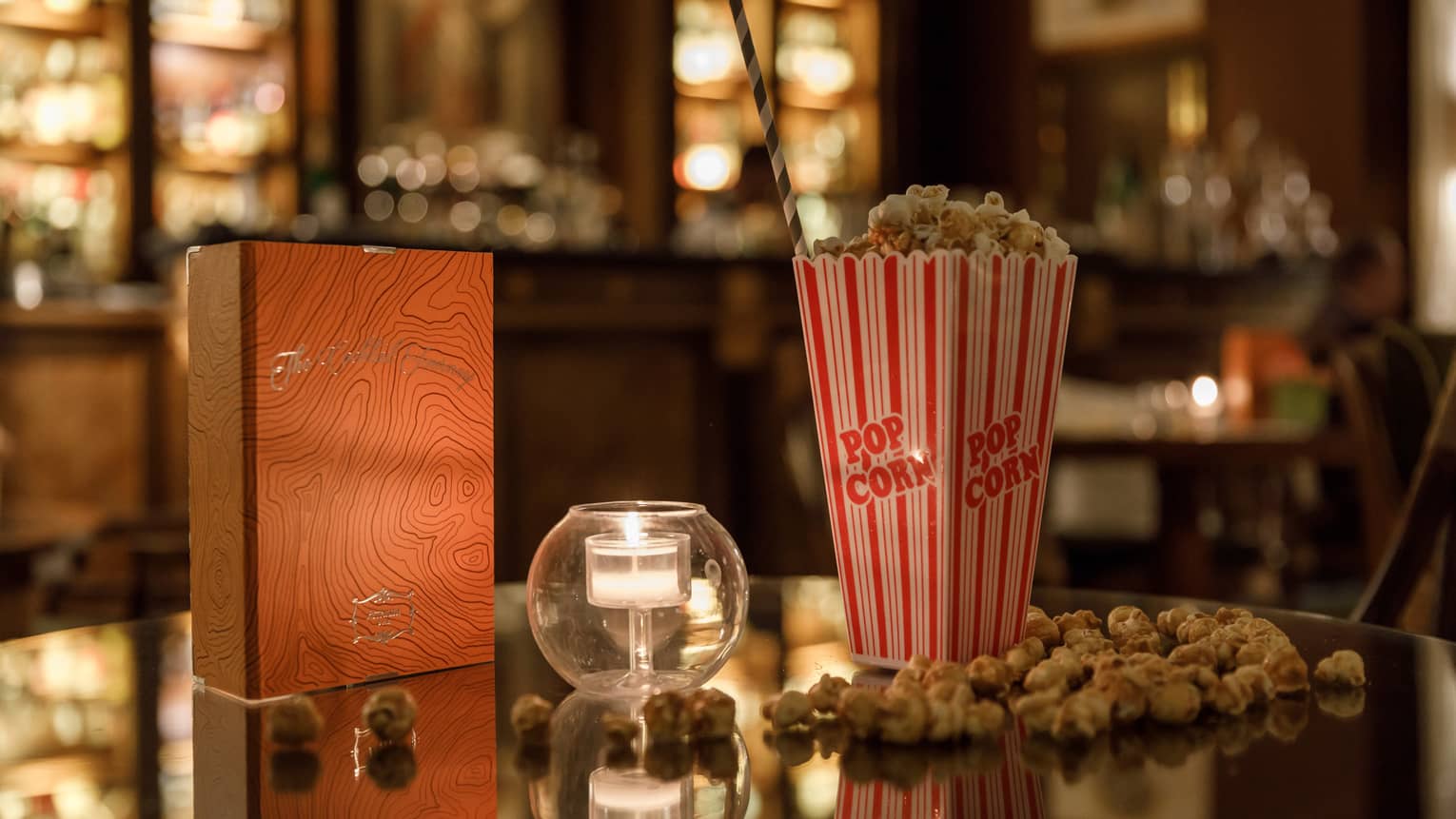 A red and gold box of chocolates, a candle in clear glass and a tall box of popcorn on a shiny table reflecting warm lights.