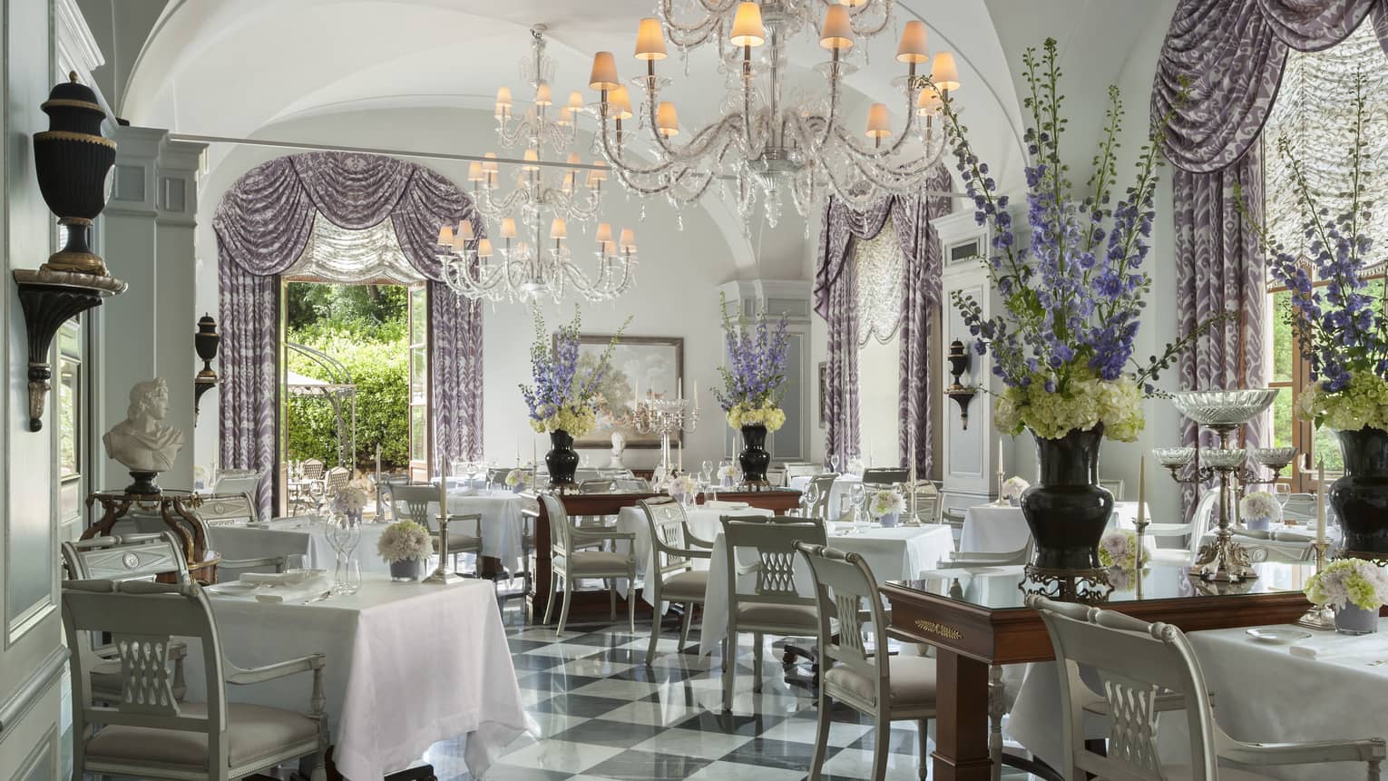 An ornately decorated dining room with warm chandelier lighting and open windows looking out on lush greenery