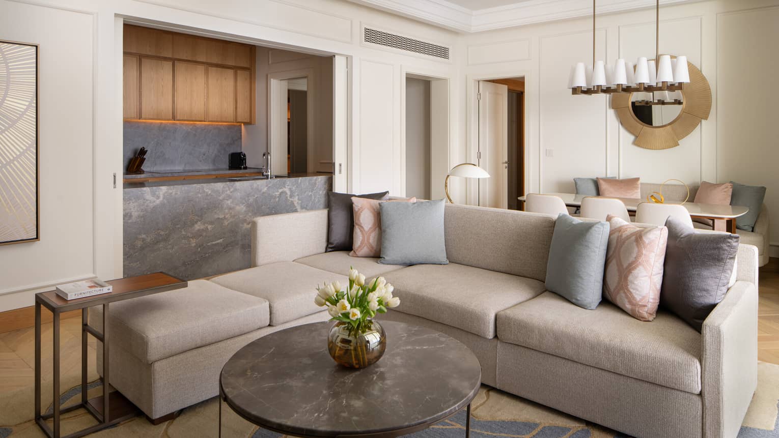 Contemporary living room with a grey sectional sofa, marble coffee table, open kitchen with wood cabinets and a dining area with pastel cushions.