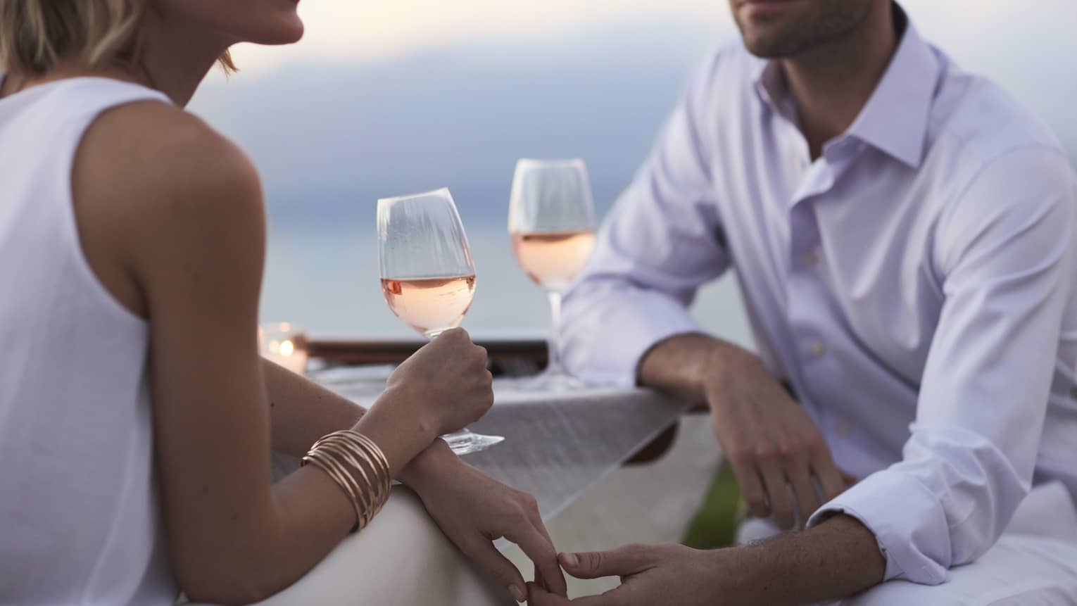 Close-up of woman and man in white, sitting and holding glasses of rose wine at dusk