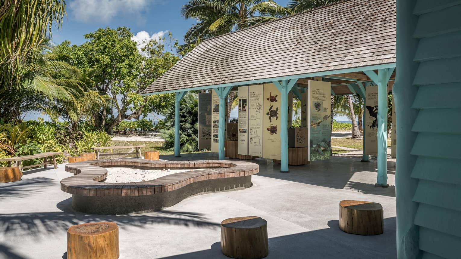 Outdoor information hut with blue poles, surrounded by palm trees, seating made of tree stumps