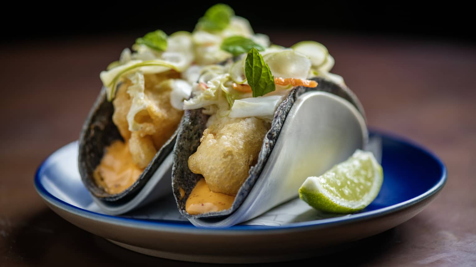 Two crispy fish tacos with aioli and cabbage-chili pickle served on a small blue plate