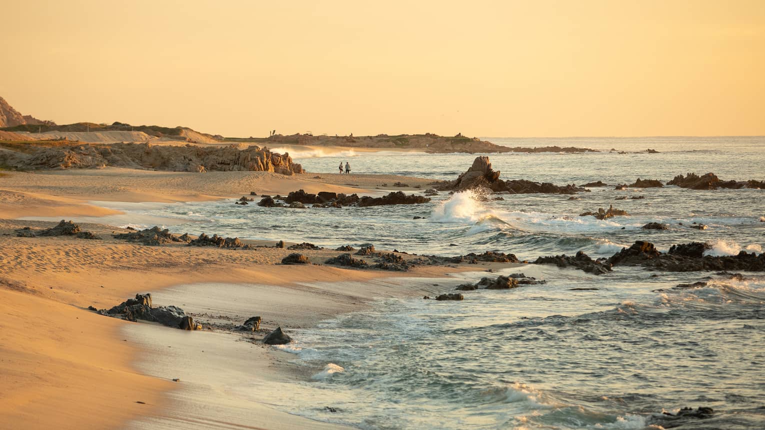 Under a clear orange sky, waves break on jutting rocks along a sandy beach as a couple standing in the distance watches.