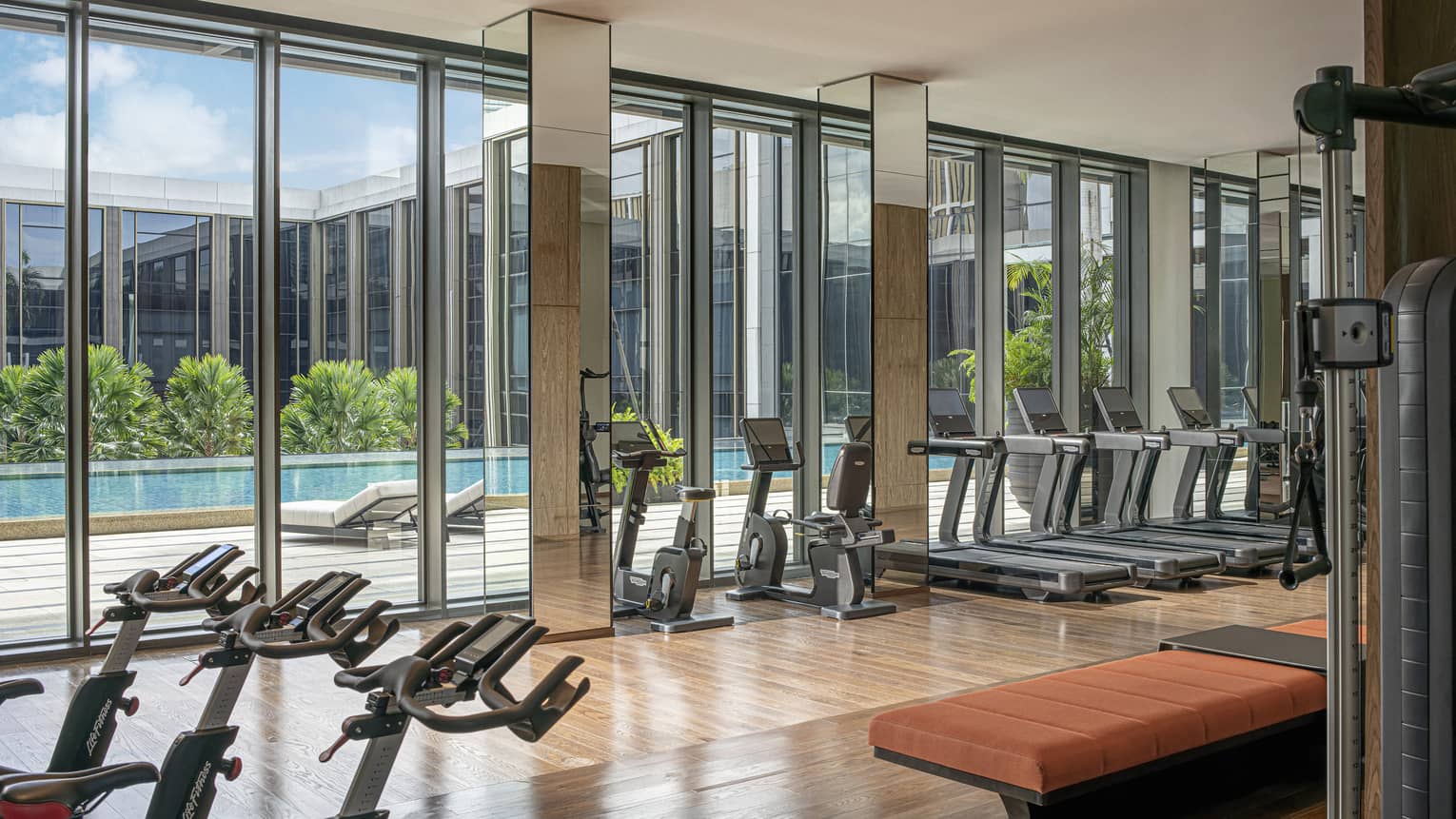 Hotel gym with floor-to-ceiling windows and rows of exercise equipment, natural light, pool views