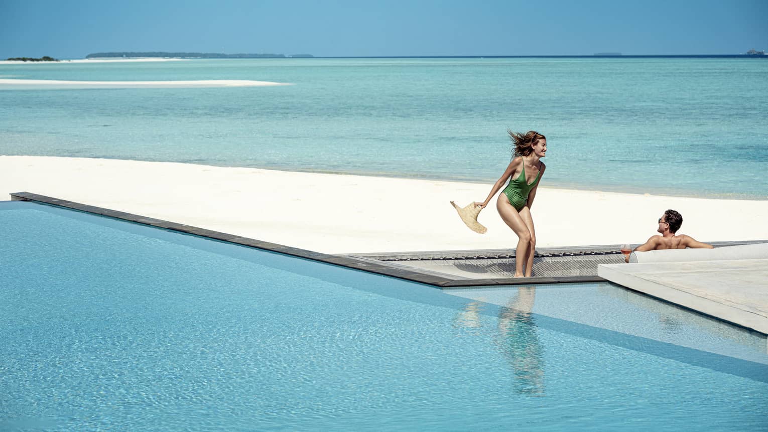 Woman and green one-piece swimsuit relaxes poolside with her male partner in the infinity pool, the vast blue waters of the lagoon behind them