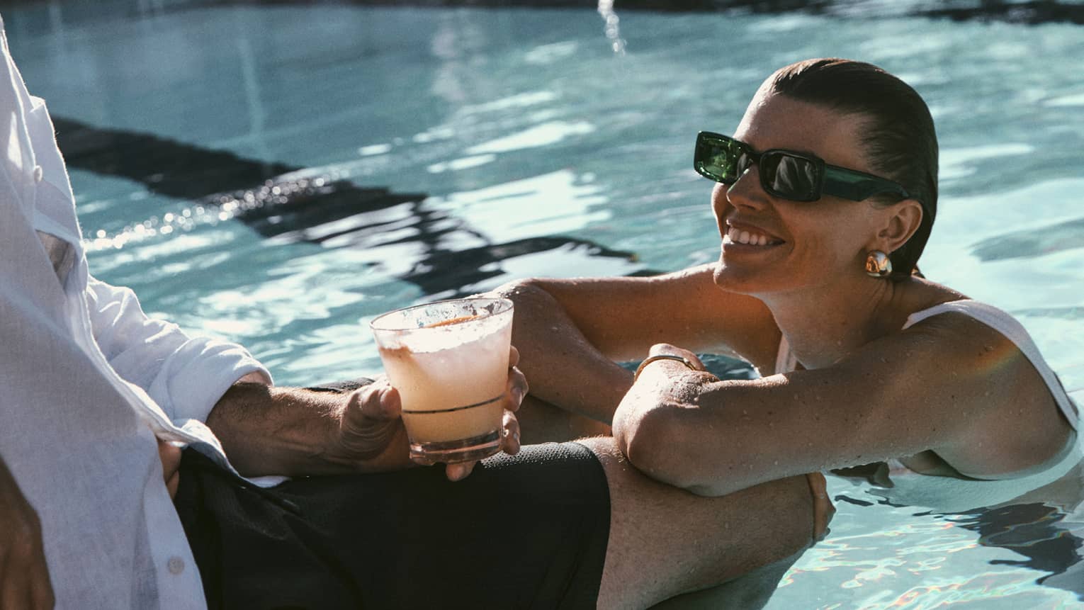 Two guests lounge by the pool, one sitting on the edge of the water holding a beverage and the other in the water