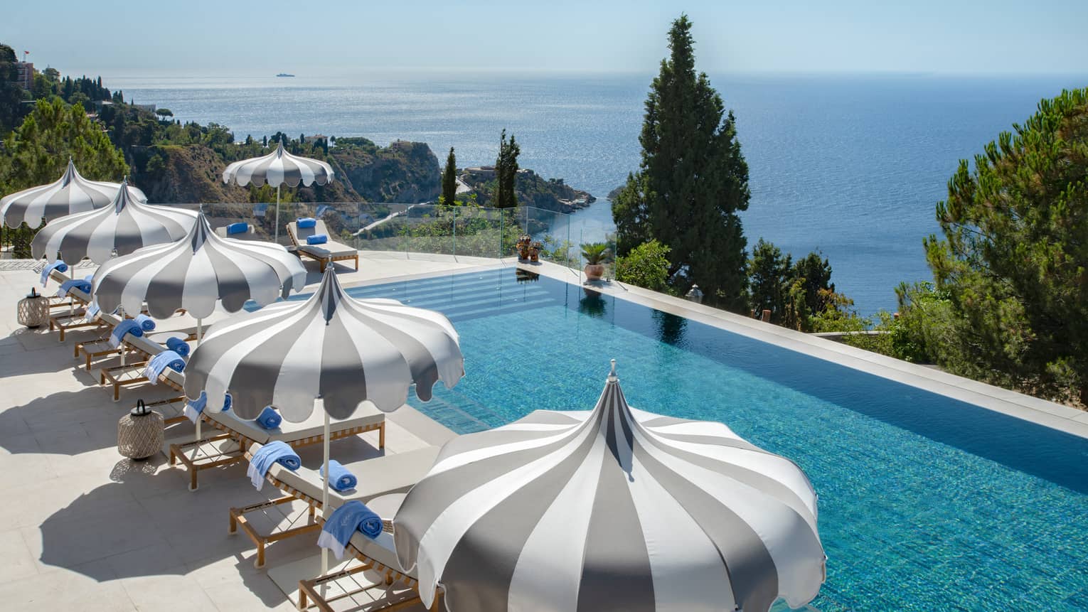 A long infinity-edge pool with lounge chairs and umbrellas along one side and one end, with trees and land and sea views in the background
