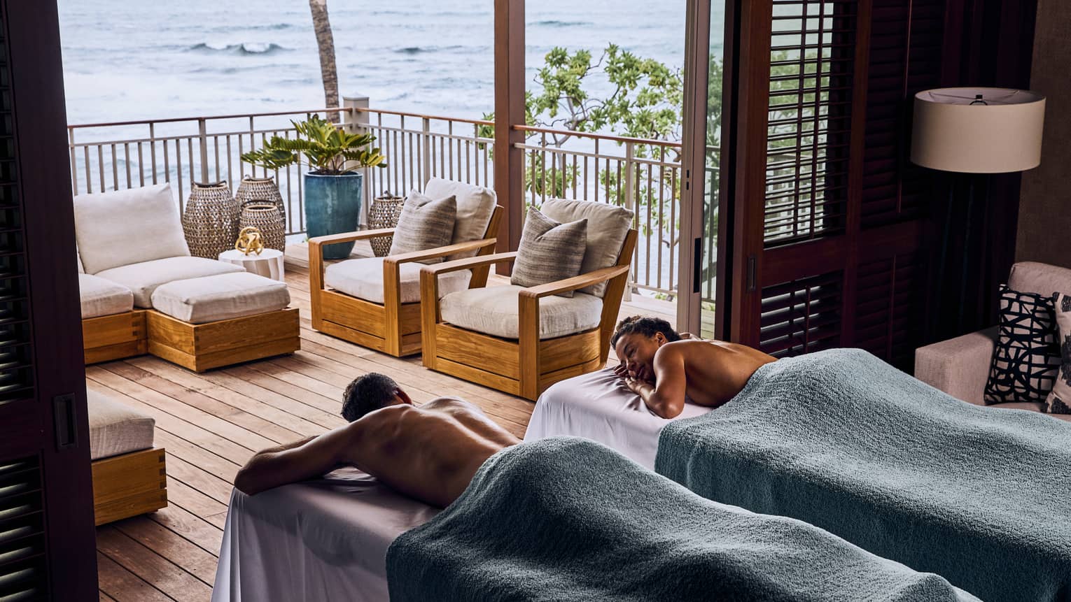 A couple covered in towels relax on massage tables in front of the open doorway of a balcony with an ocean view.