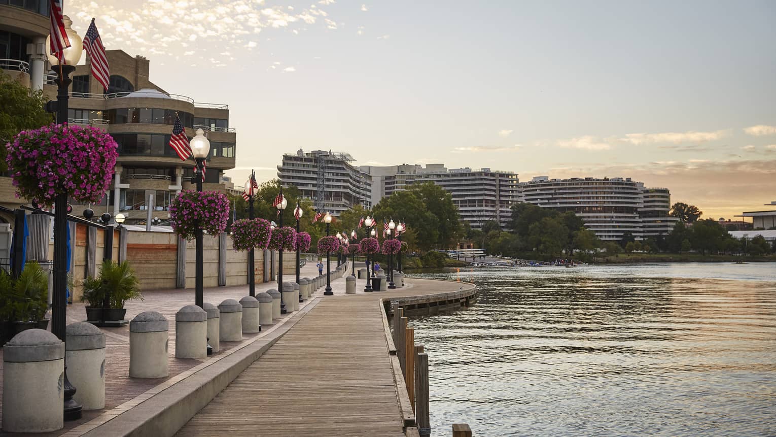 A waterfront with a wooden walkway next to light posts and flowers with building on the other side of a small road.