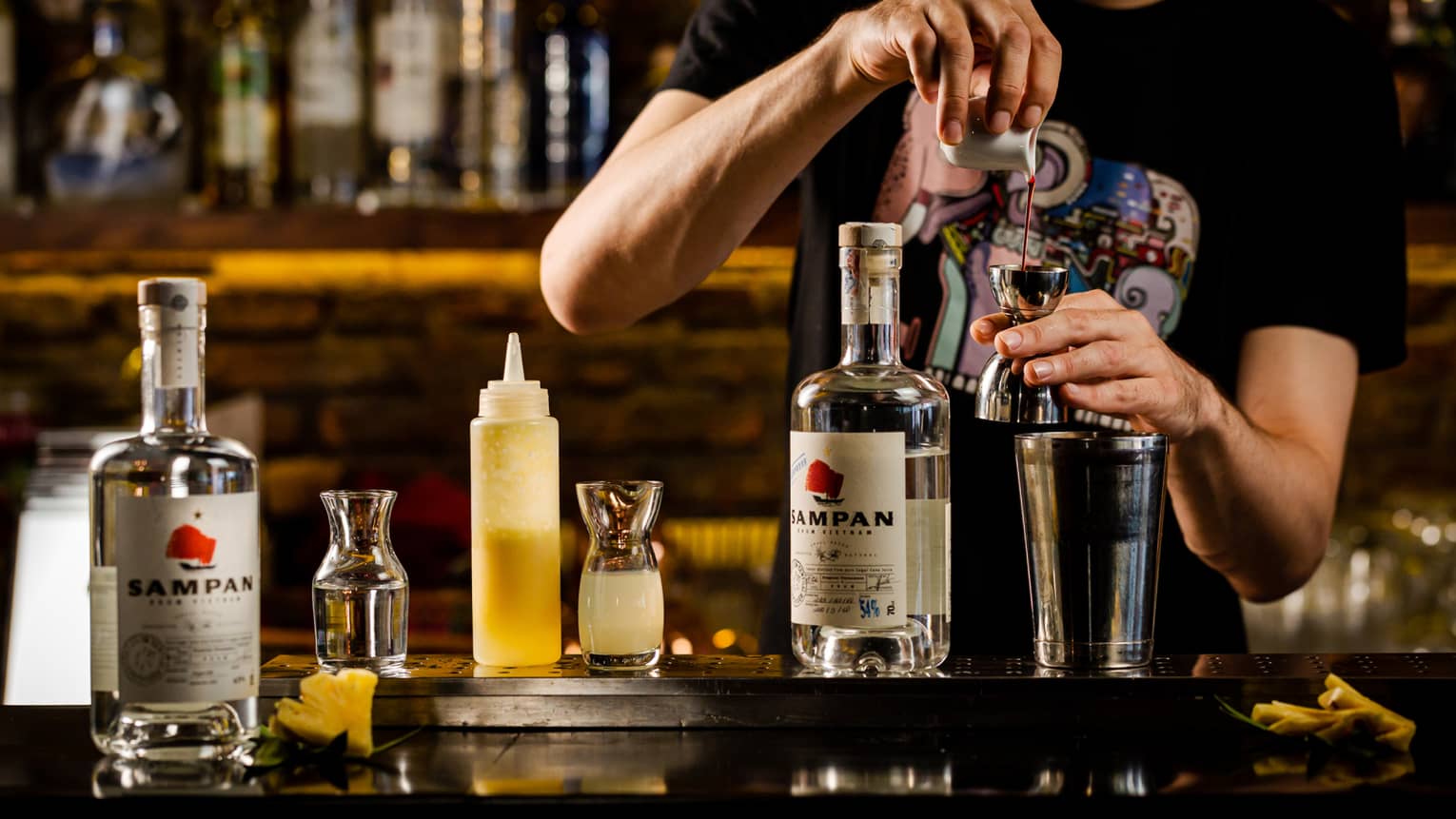 Bartender pours Sampan rhum into cocktail shaker, bottles of the liquor displayed on bartop
