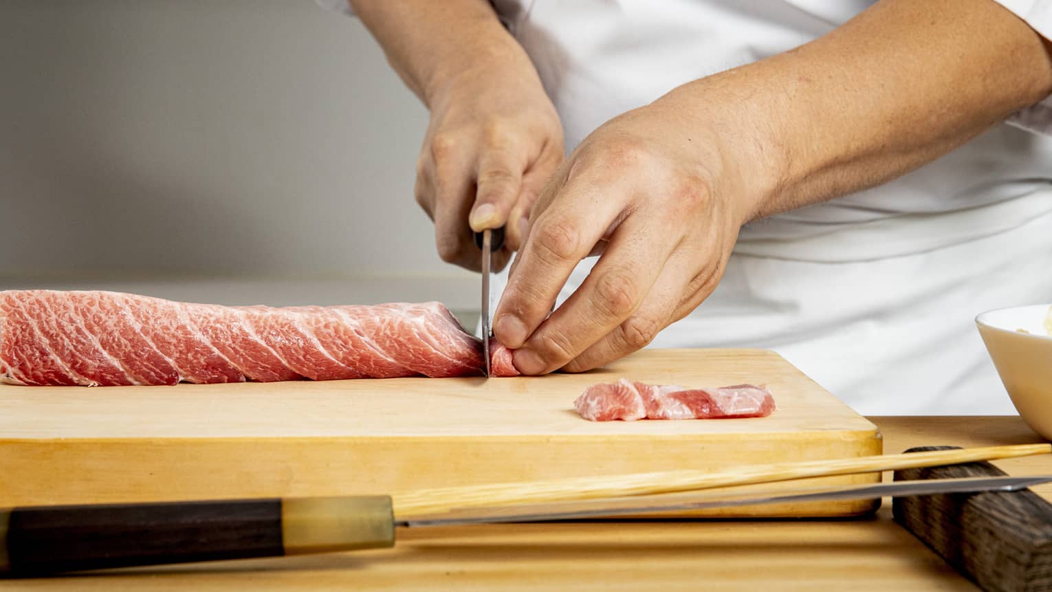 Chef's hands use knife to thinly slice meat on wooden board, preparing it for sushi