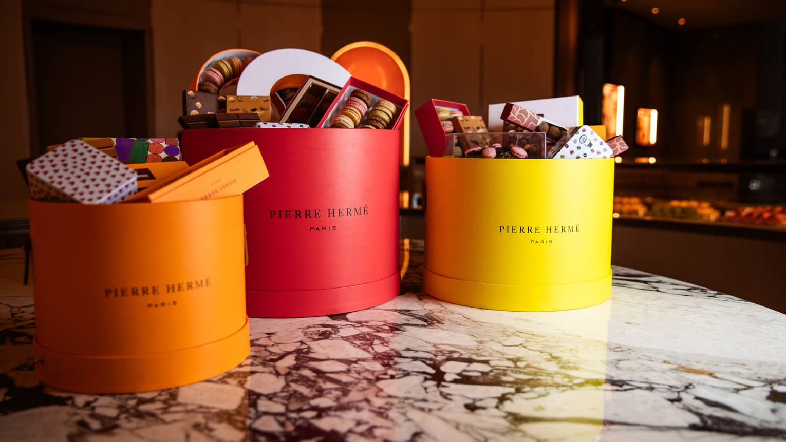 Three cylyndrical gift boxes ? one orange, one red and one yellow ? sit filled with desserts and goodies on a white-and-black marble table