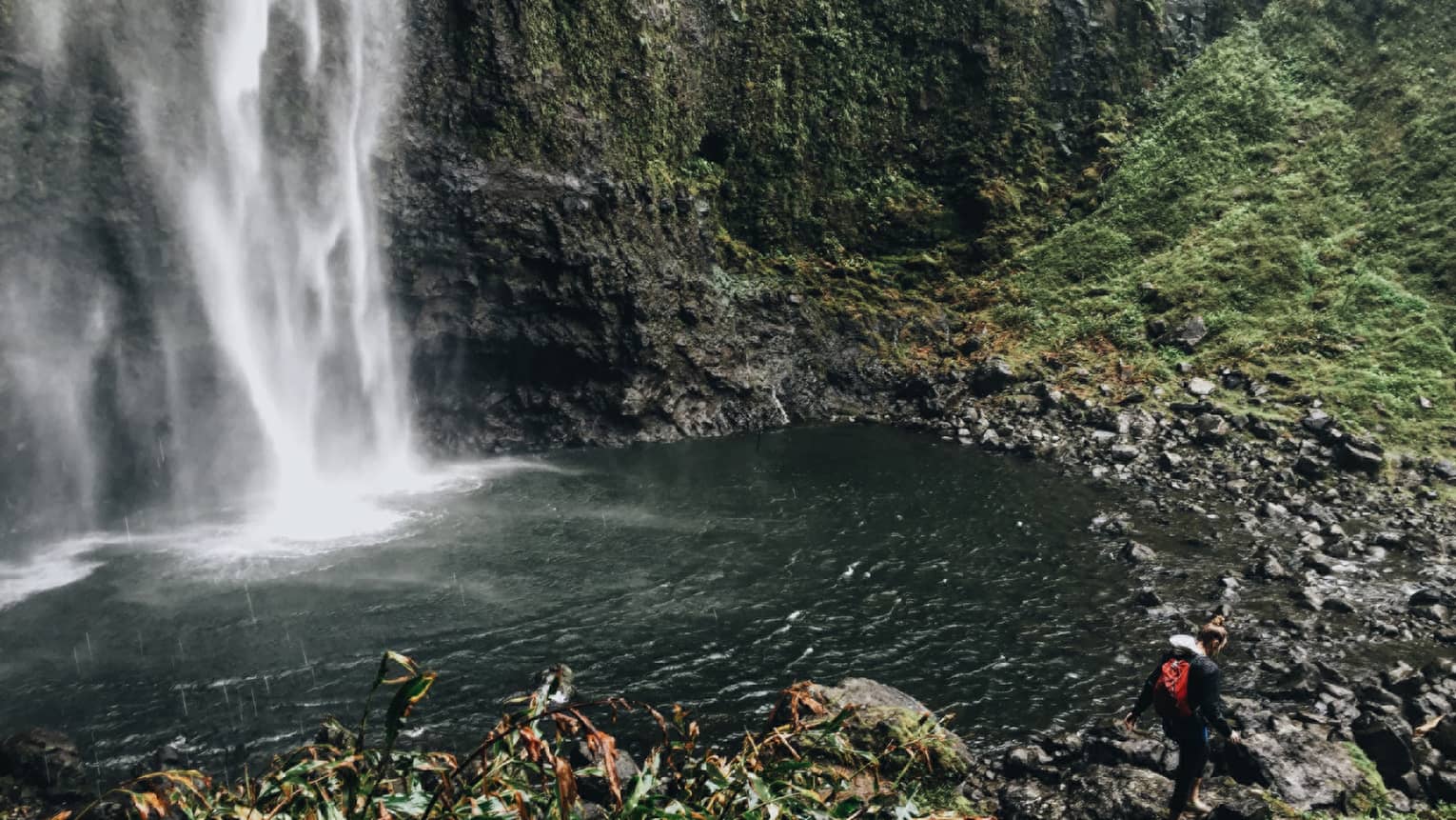 Back of woman hiking over black rocks under tall waterfall, small pond
