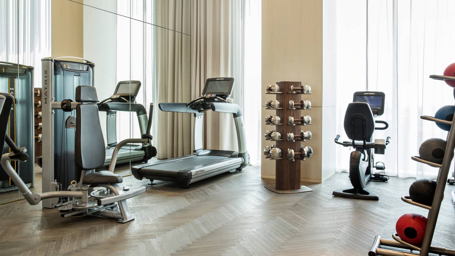 Cardio exercise machines, rack of hand weights near windows with long curtains in fitness centre