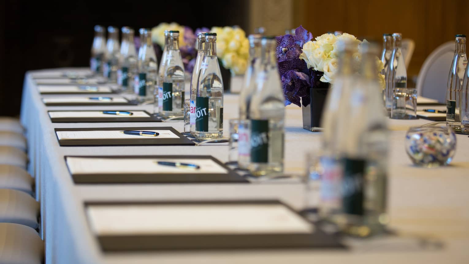 Row of meeting agendas, glass bottles, small vases with flowers on boardroom table