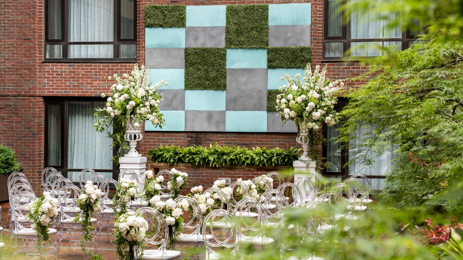 An outdoor area with rows of white seats and flowers arranged around them, a brick walled building can be seen in front of the chairs.