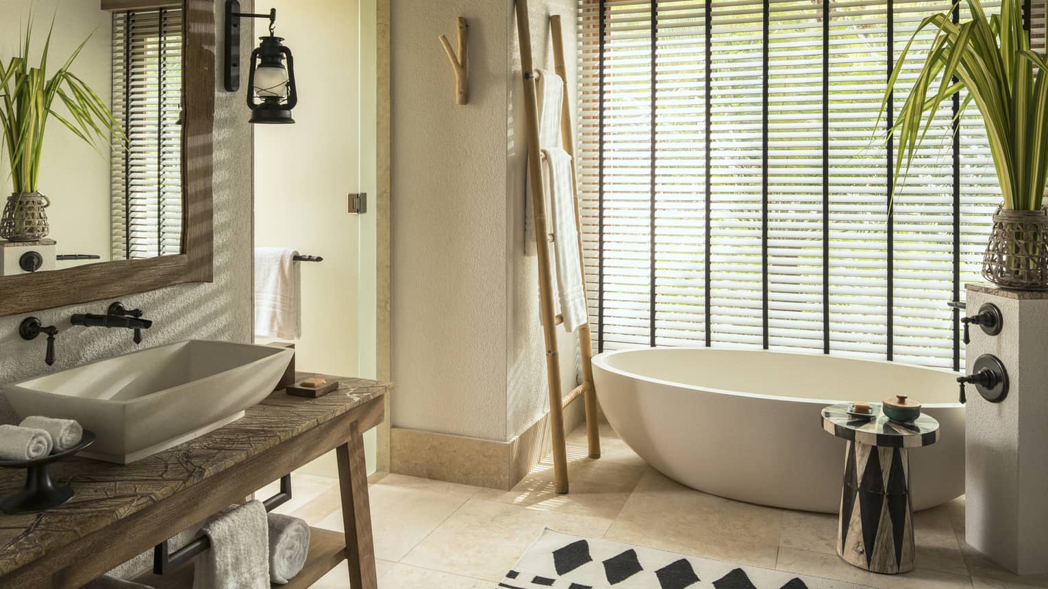 Free-standing white tub in bathroom under window with closed blinds, sink and vanity