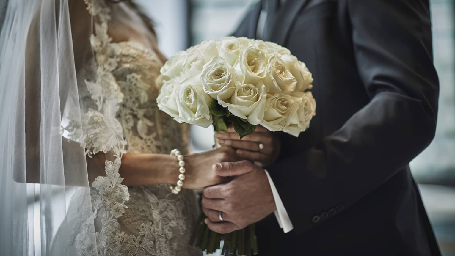 A close up of a bride in a lace gown and a groom in a gray suit hold the bride's bouquet of white roses together.