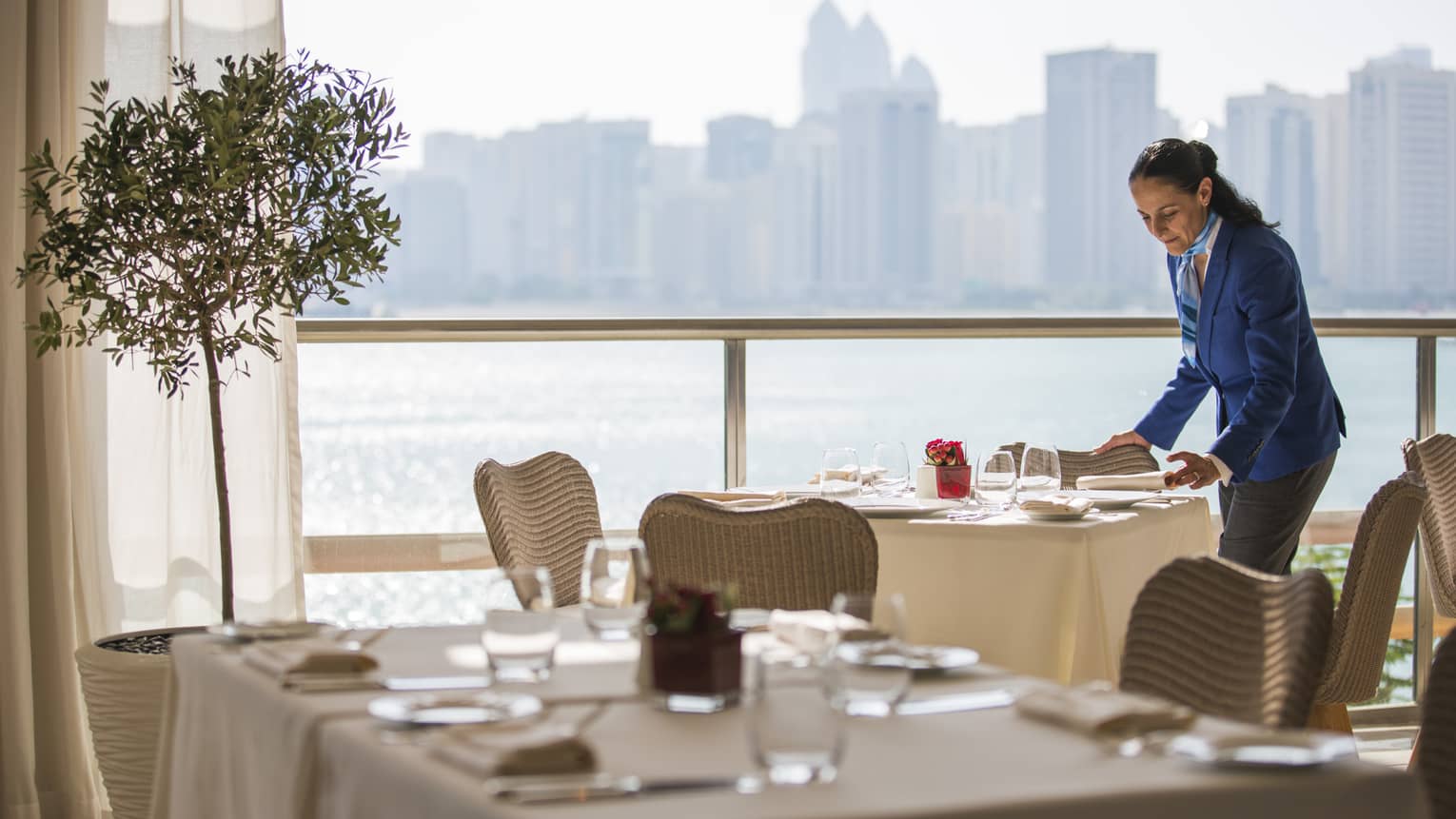 Woman in blue suit sets table by sunny window with water views at Cafe Milano