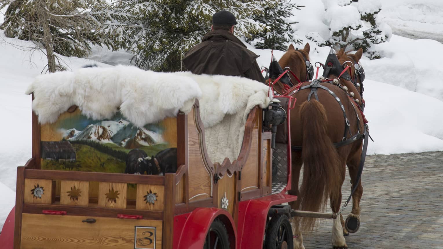 Rear view of a painted horse-drawn carriage piled with furs travelling towards a cabin through a snowy, evergreen forest.