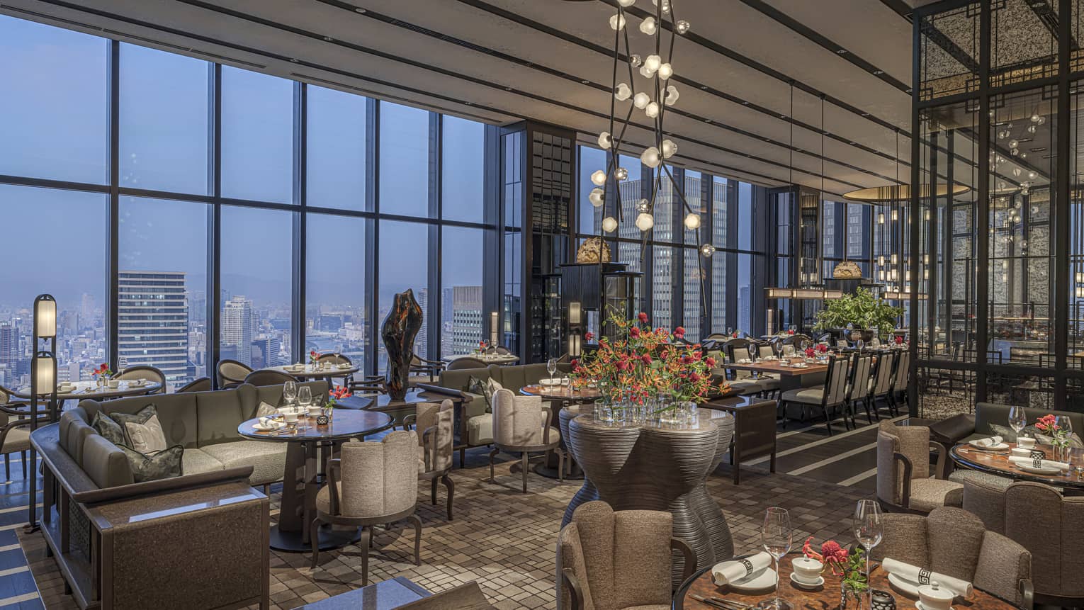 Expansive high-end restaurant warmly lit by modern chandeliers with a bank of windows offering aerial views of the city