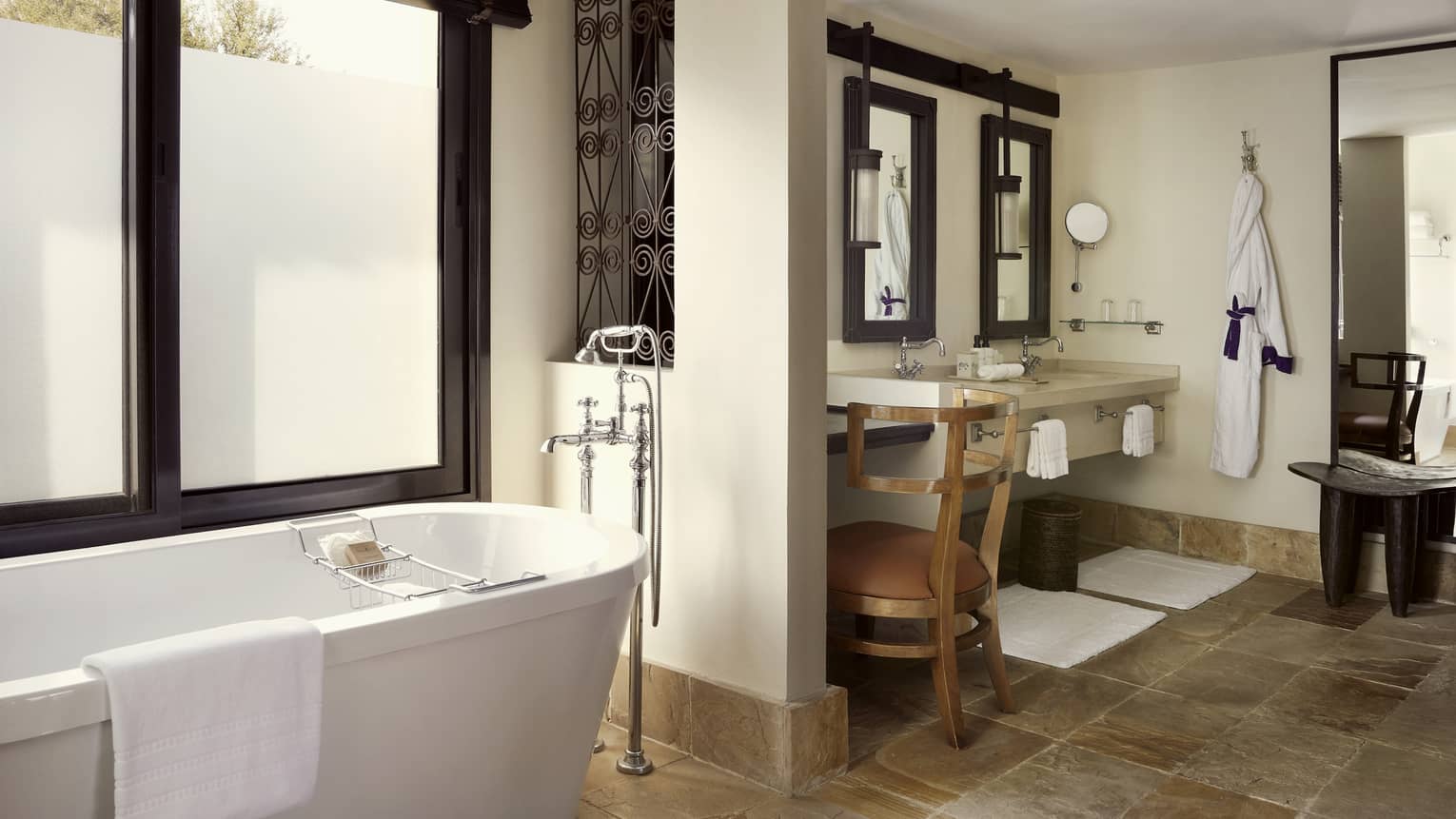 Bathroom with deep soaking tub against window and double vanity under rustic mirrors with brown desk chair, all on slate flooring