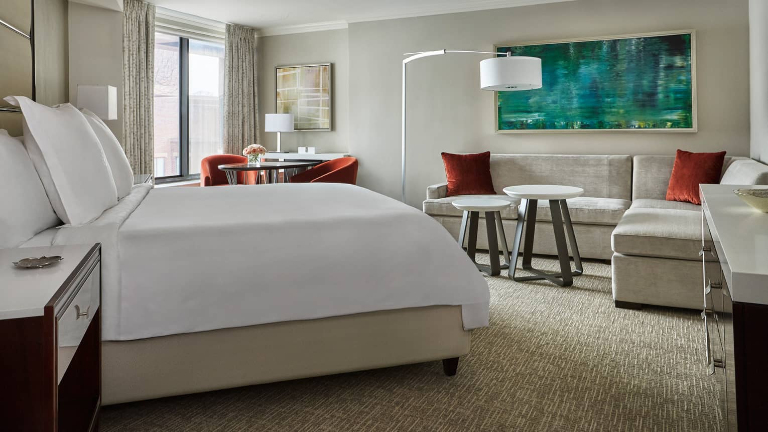Deluxe Room side of bed with white linens across from L-shaped grey sofa with red pillows, table by window