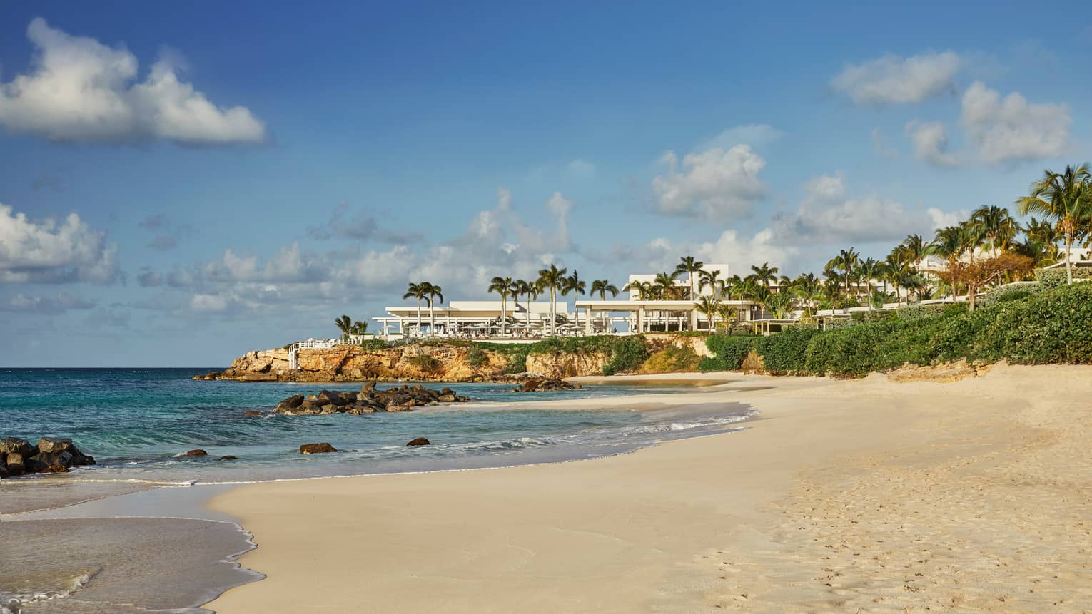 Sandy beach with turquoise water and a luxury resort nestled on a palm-lined cliff.