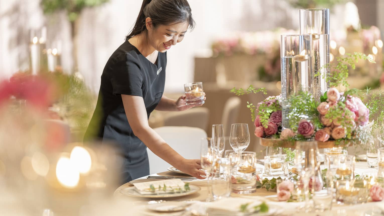 A smiling staff member arranging a table with a floral centerpiece, long glass jars with floating tea lights and champagne glasses