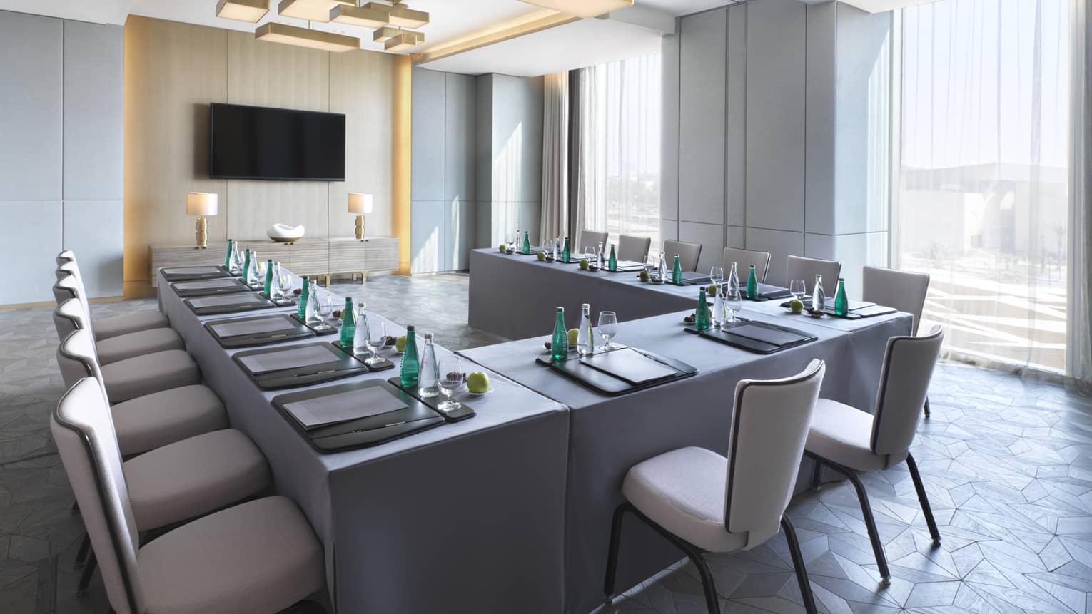 U-shaped desks with water, stationery and fruit in light-filled meeting room facing TV screen