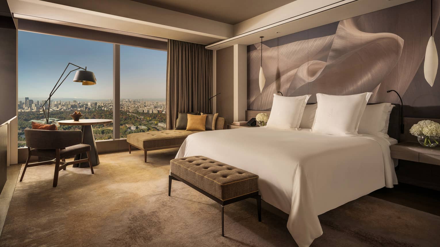 Hotel room with a cushioned bench at the end of white king bed and a wall of windows