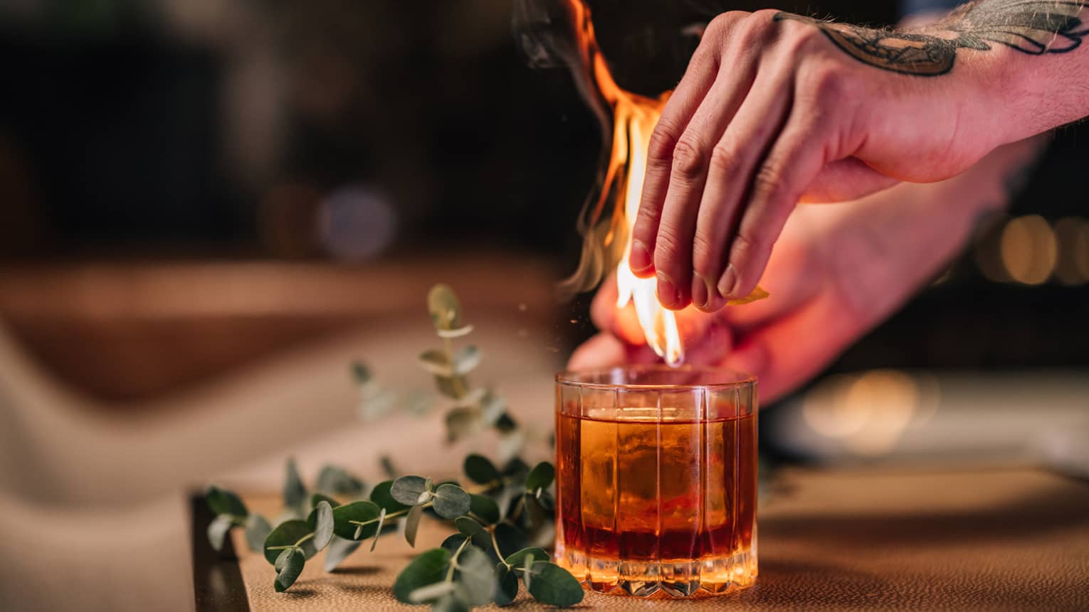 Bartender's tattooed hands light a flame above a cocktail served in a rocks glass next to a sprig of fresh eucalyptus