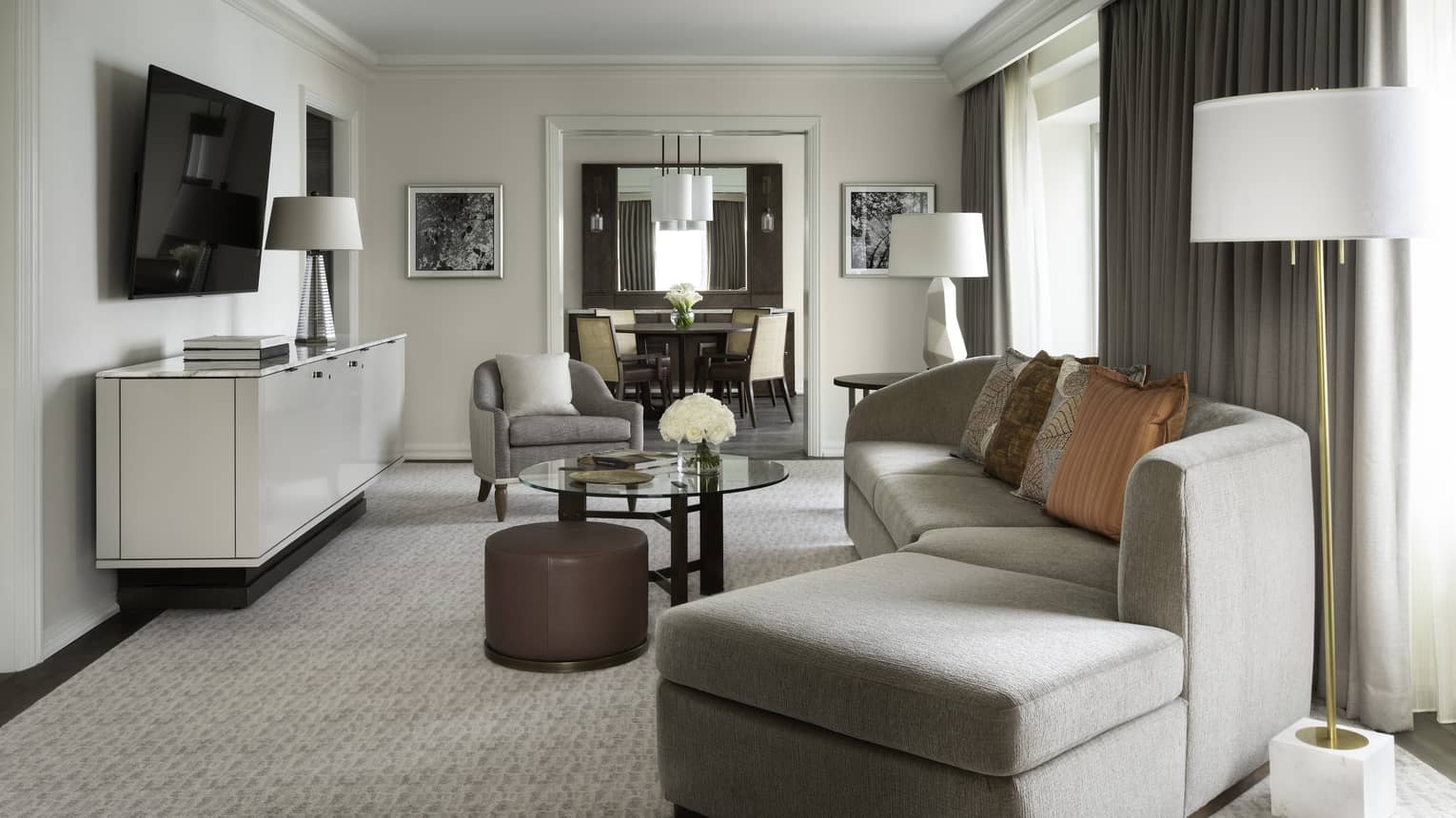 Living area with grey curved sofa, glass table, beige carpet, TV on wall, curtains