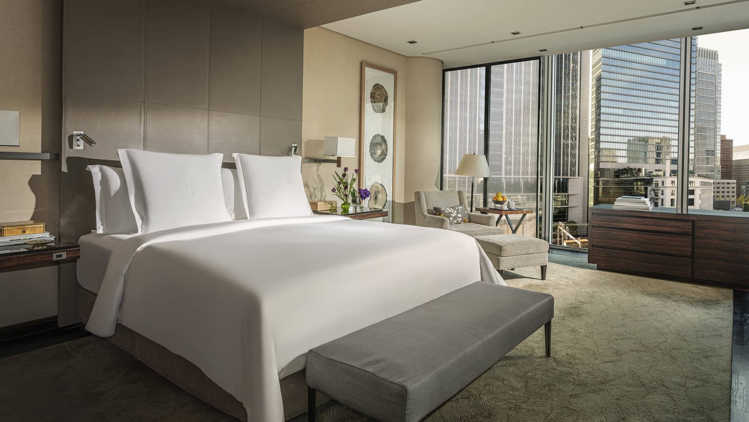 Luxury hotel room with a king bed, armchair, floor-to-ceiling windows with city skyline views and wooden dresser
