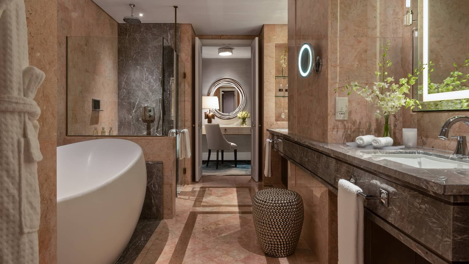 Bathroom with marble walls, featuring a freestanding white bathtub, glass-enclosed shower, double vanity and seating area with a circular mirror.