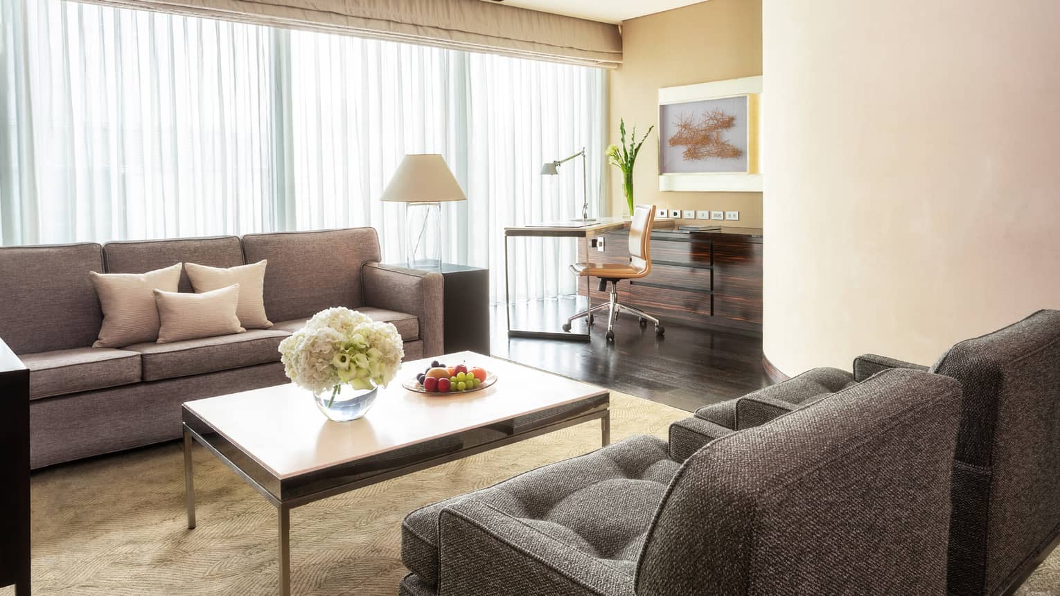 Living area with grey sofas, coffee table with flowers and fruit, work desk with a chair and floor-to-ceiling windows with curtains