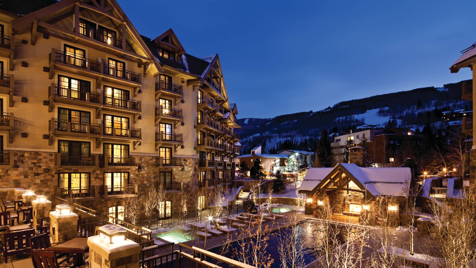 Mountain resort at night with illuminated balconies, outdoor hot tubs, and snow-covered surroundings.
