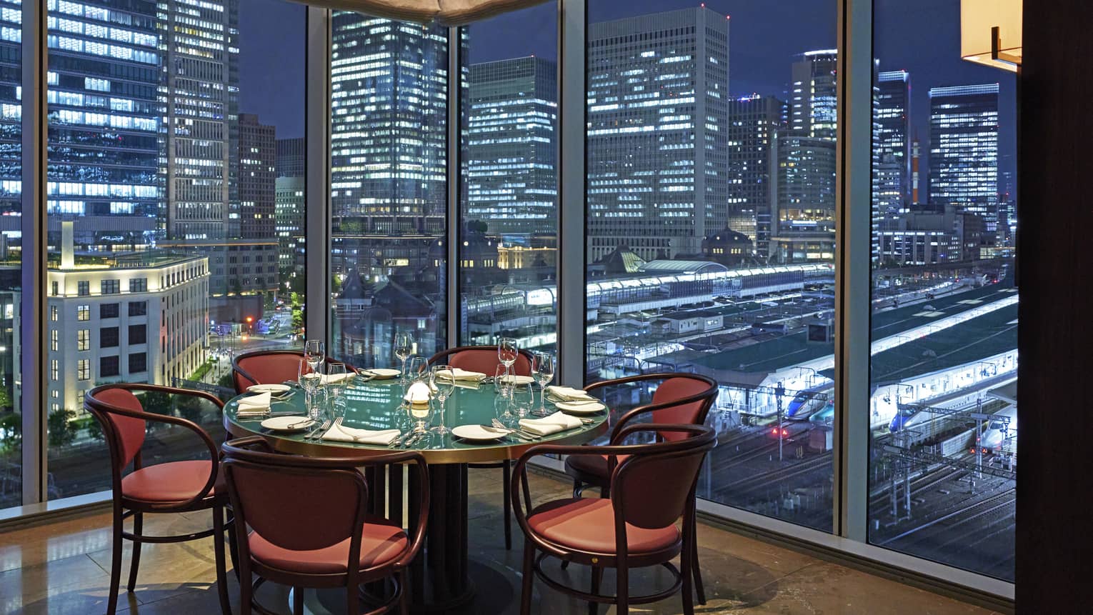 Luxury boutique hotel dining setup with a round table elegantly set for guests, offering a panoramic city view at night through tall glass windows